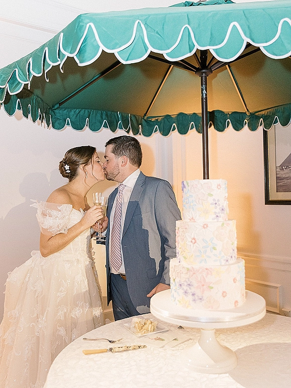 Wedding kiss as the couple leans together by a three-tier cake cutting moment, champagne flute in hand, under a teal umbrella indoors