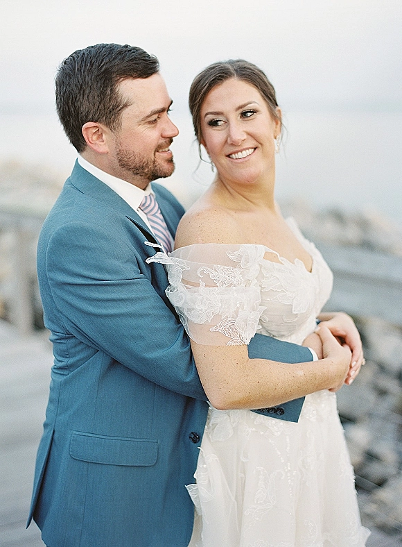 Couple portrait of bride and groom embrace on a rocky shoreline with ocean and sky, her off-the-shoulder lace dress visible as he hugs her