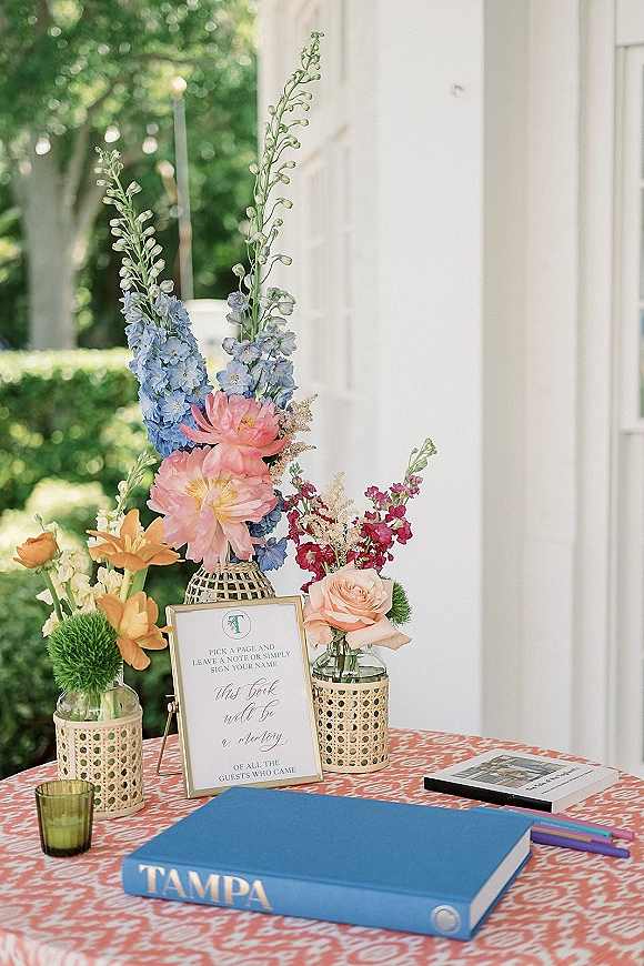 Wedding guest book table with a guest book, framed sign, colorful pens, and bright floral vases on a patterned cloth by a white wall window