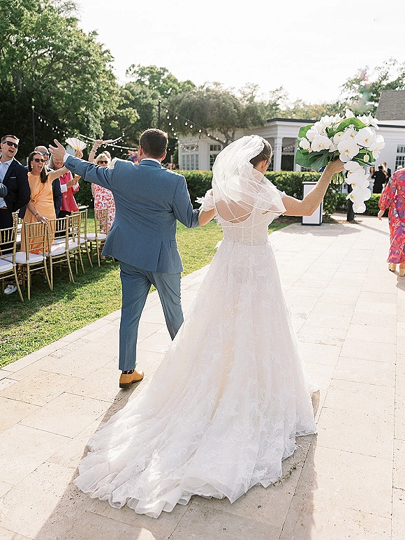 Wedding recessional as bride and groom walk away cheering, bride lifting white orchid bouquet with cathedral veil along garden aisle walkway