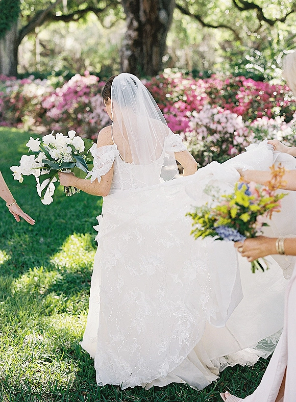 Bride portrait from behind as she walks on grass holding a white and green bouquet, long veil and off-the-shoulder dress in a garden lawn setting