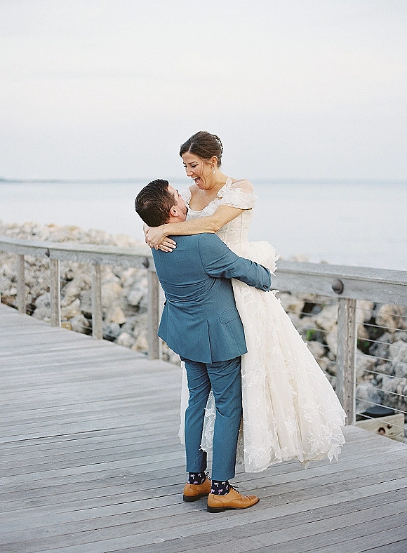 Couple portrait of groom lifting laughing bride in an off-the-shoulder lace gown on a wooden boardwalk with ocean and sky behind