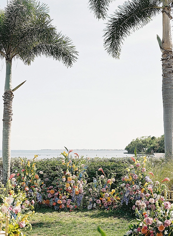 Ceremony florals and wedding aisle florals in low clusters of roses, greenery, and ornamental grass on a lawn with palm trees and ocean view