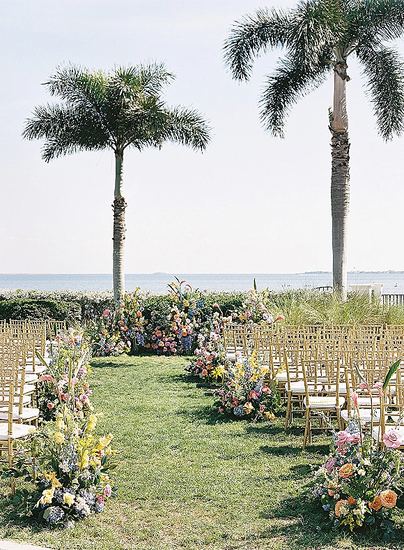 Ceremony setup with floral aisle arrangements and gold chiavari chairs on a lawn overlooking the ocean with palm trees and sky