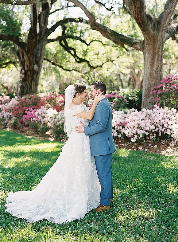Couple portrait of bride and groom embrace in a garden, bride in lace gown and veil holding his face beneath oak trees