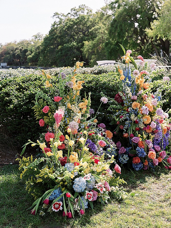 Ceremony floral arrangements of colorful garden roses clustered with wildflowers along a lawn aisle, set before boxwood hedges in daylight