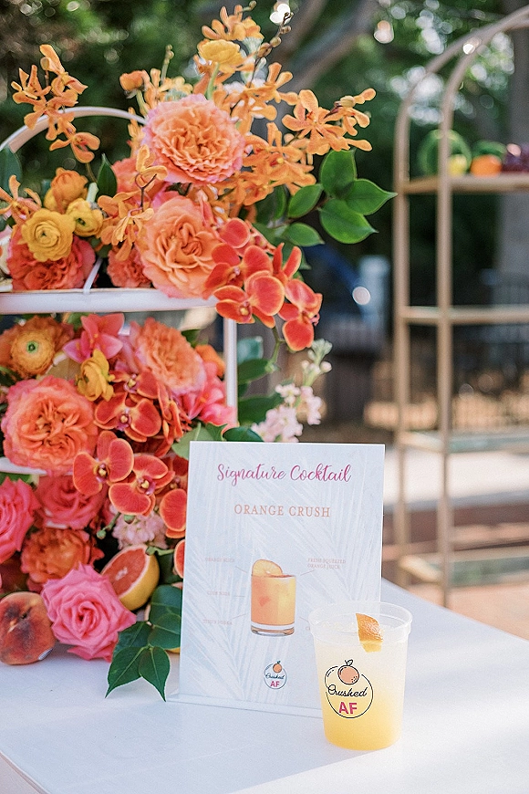 Signature cocktail sign with a wedding signature drink sign on a white shelf, framed by coral roses and orchids against greenery bokeh lights