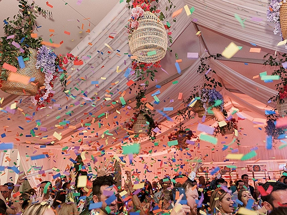Wedding reception party with a confetti toss as guests cheer with hands raised under draped tent ceiling, wicker lanterns and flowers