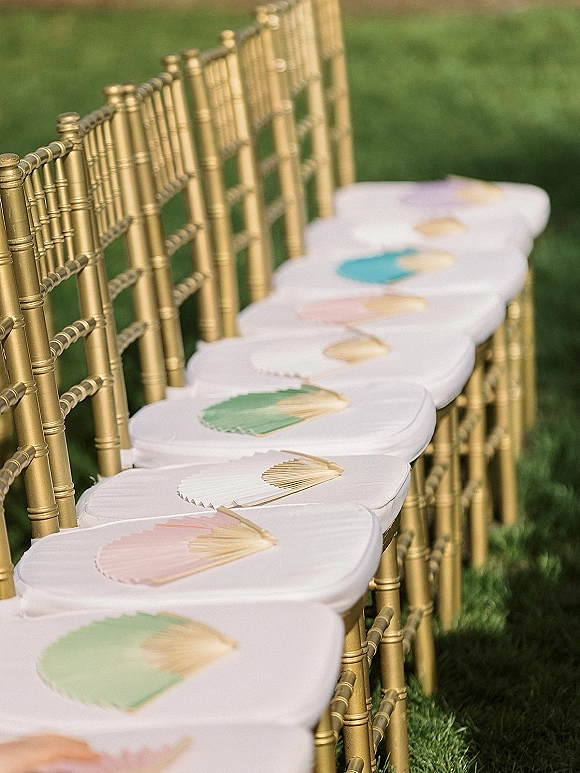 Ceremony seating with gold chiavari chairs lined on a green grass lawn, each with white cushions and a handheld paper fan on the seat