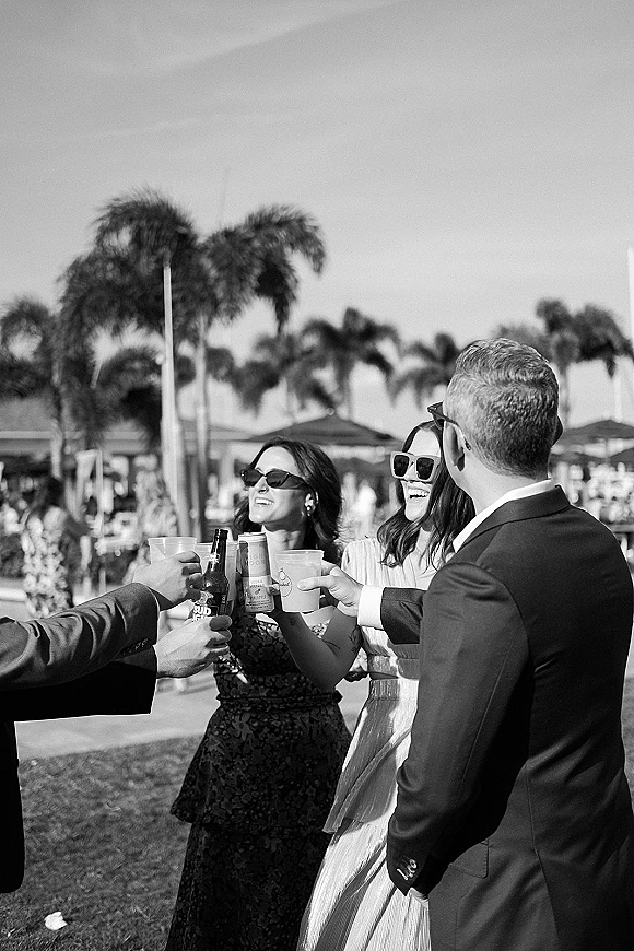 Wedding reception toast as guests cheer with plastic cups and a beer bottle, one in sunglasses, on a palm-lined resort lawn under blue sky