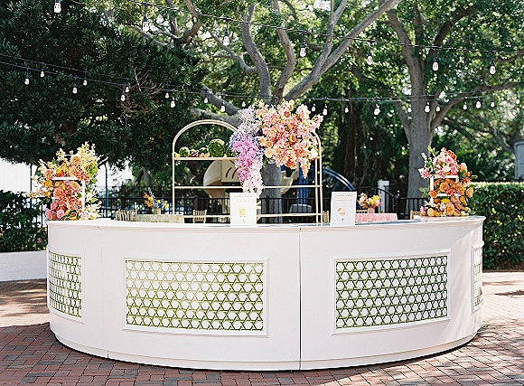 Wedding bar setup with a curved white bar, pink and peach florals, and citrus accents under string lights on a brick garden patio