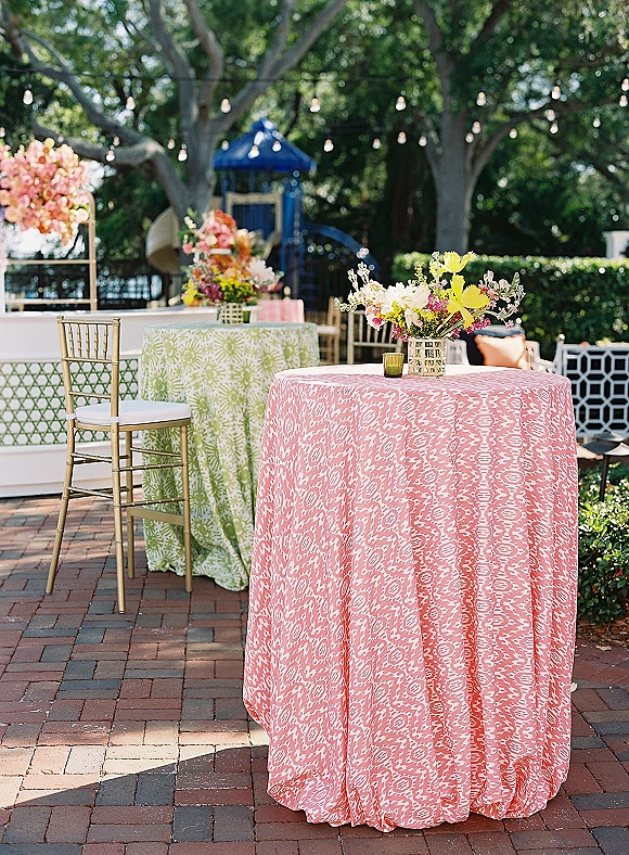 Cocktail hour lounge with outdoor cocktail tables in pink ikat linens, gold Chiavari chairs, and floral centerpieces under string lights on a brick patio