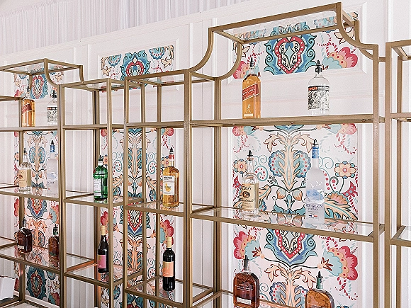 Wedding bar display with brass shelving and glass shelves holding liquor and wine bottles against patterned wallpaper and white paneling
