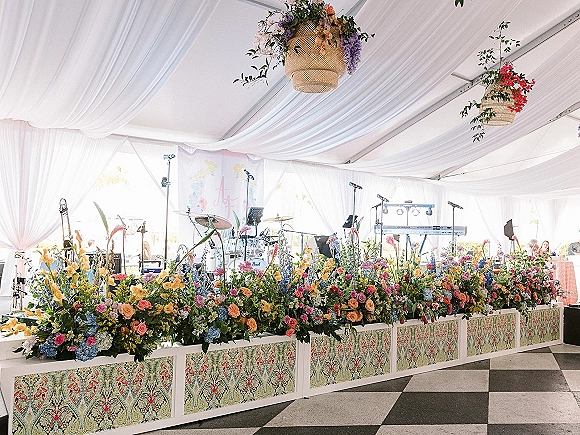 Wedding band stage with floral border and draped tent ceiling, featuring drum kit, keyboard, and hanging wicker lanterns above dance floor
