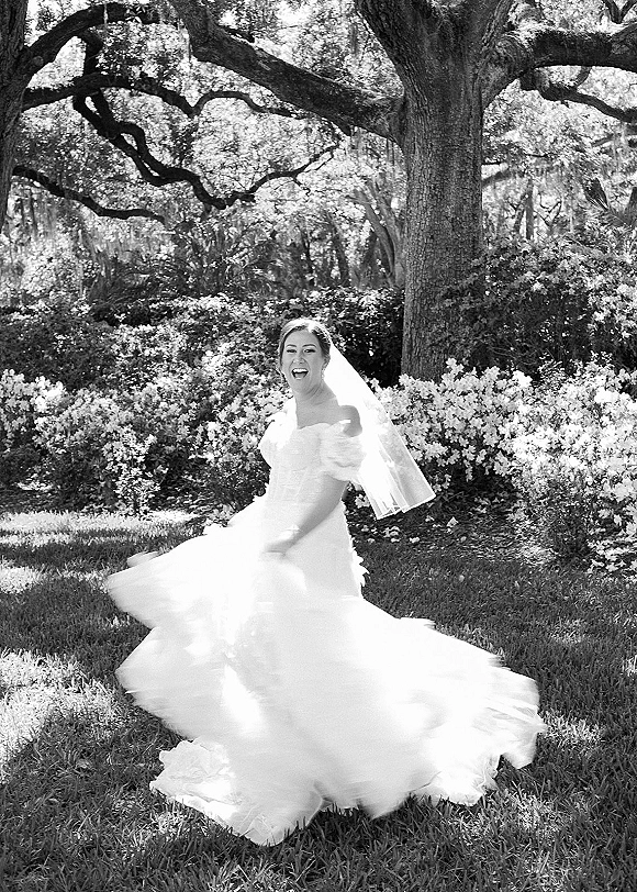 Bridal portrait of a bride twirling her wedding dress with veil flowing, laughing on a garden lawn with trees and flowering bushes
