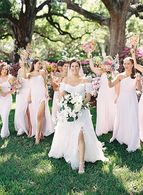 Bride with bridesmaids walking together in pastel dresses, holding colorful bouquets, with a veil trailing in a lush garden setting