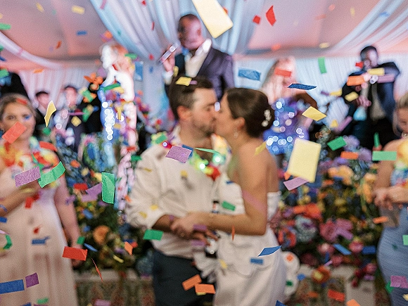 Wedding kiss as colorful confetti falls around the bride and groom on a reception stage under string lights and a draped tent ceiling