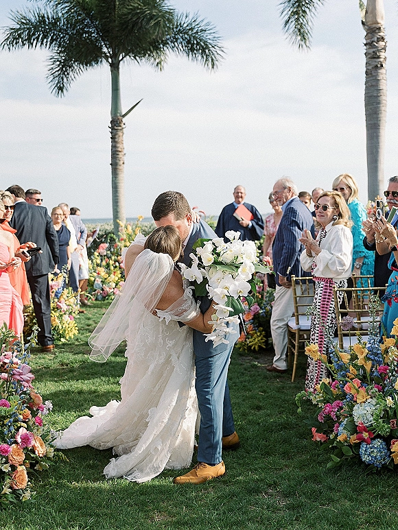Wedding kiss as bride in veil and dress kisses groom in suit, holding orchid bouquet on a palm-lined oceanfront aisle with guests behind