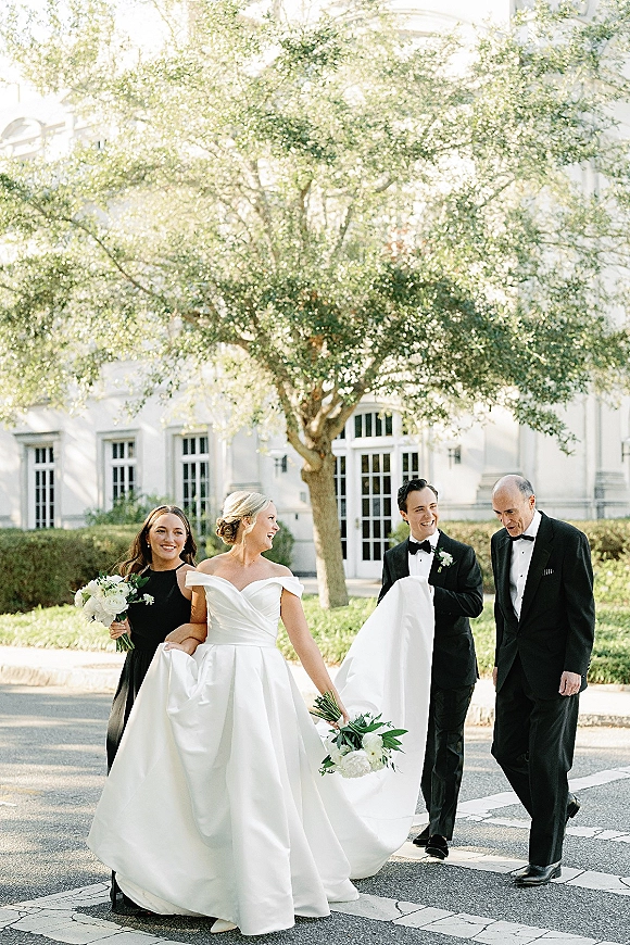 Wedding party portrait of bride and groom walking with bridesmaids and groomsmen, bride holding bouquet near a white building on lawn walkway