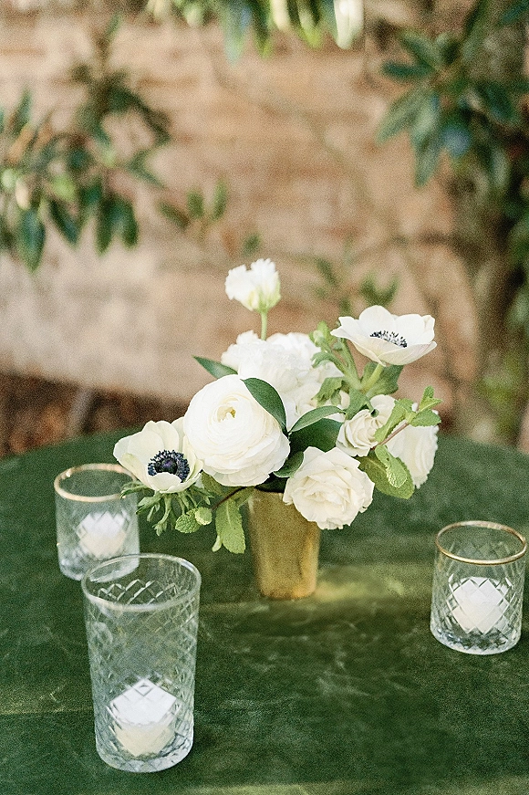 Wedding centerpiece with white flowers and greenery in a brass vase, surrounded by cut glass candle holders on a wood table by a stone wall