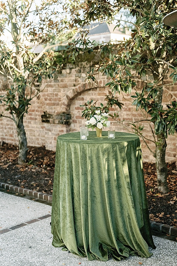 Cocktail table decor on a green velvet tablecloth with a white floral centerpiece in a gold vase and glass votive candles by a brick wall patio