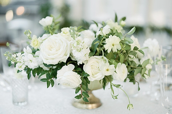 Wedding centerpiece with a white rose centerpiece of lush greenery in a low gold compote vase on a white tablecloth in soft indoor light