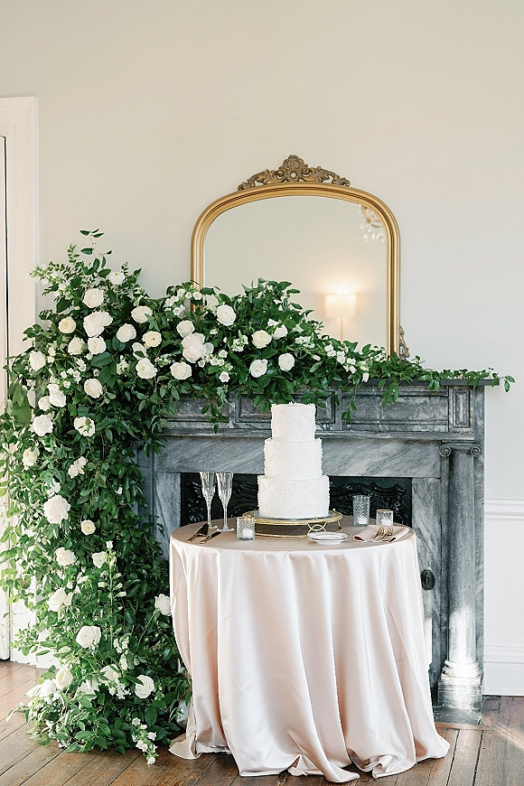 Wedding cake table with a three tier wedding cake on a stand, blush linen and candles, set by a marble fireplace and gold mirror
