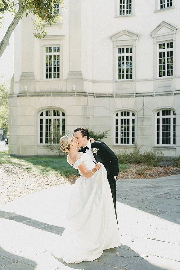 Wedding kiss portrait of bride and groom kissing in a dip, off-the-shoulder gown and tuxedo, by a white building with tall windows