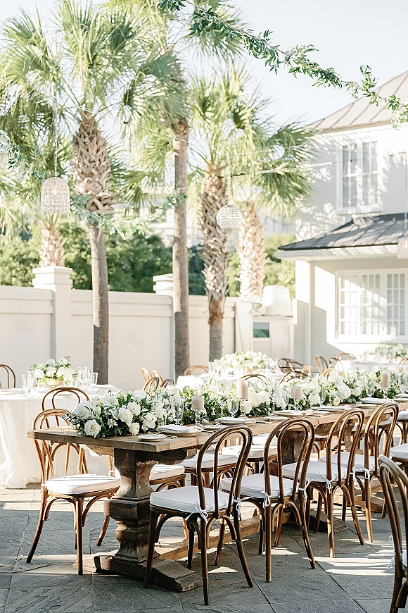Reception tablescape with an outdoor wedding reception table on a wood farm table, white roses and greenery garland under string lights on a patio with palms