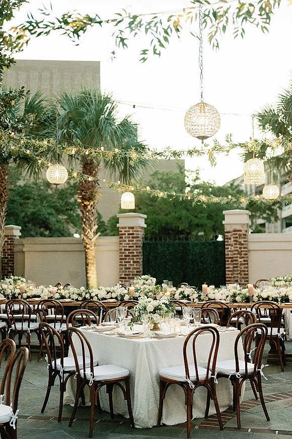 Outdoor reception decor with round and banquet tables in white linens, floral centerpieces and candles under cafe string lights in a courtyard patio