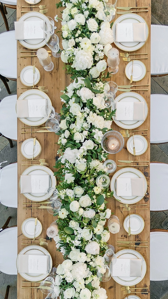 Reception tablescape with long wedding table setup on a wood dining table, white rose and greenery garland, gold flatware, taper candles on a stone patio