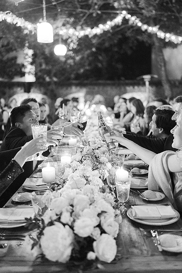 Wedding reception toast as guests clink wine glasses and a beer bottle over a candlelit long wooden banquet table under string lights outdoors
