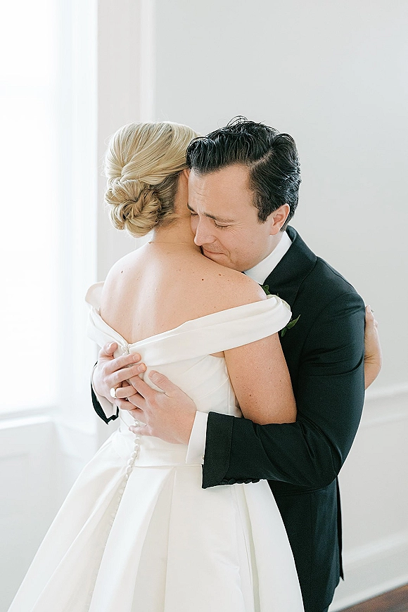 Wedding couple portrait of bride and groom hug, forehead-to-forehead as he embraces her off-the-shoulder dress in soft window light by a white wall