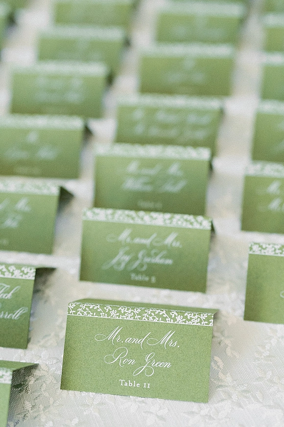 Wedding place cards in green with calligraphy lettering and floral border pattern on a white lace tablecloth beside table numbers