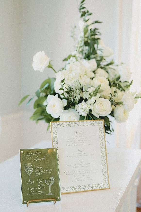 Wedding bar signage with signature drinks sign on a stand, paired with white roses, hydrangea, and greenery on a white tablecloth