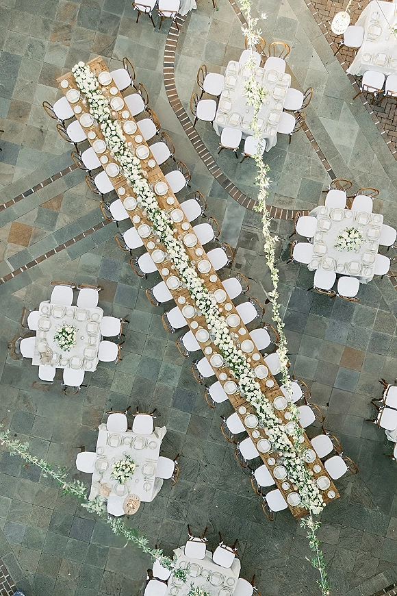 Reception tablescape with long banquet table decor, white linens, floral garlands and wicker lanterns on a stone patio with brick border