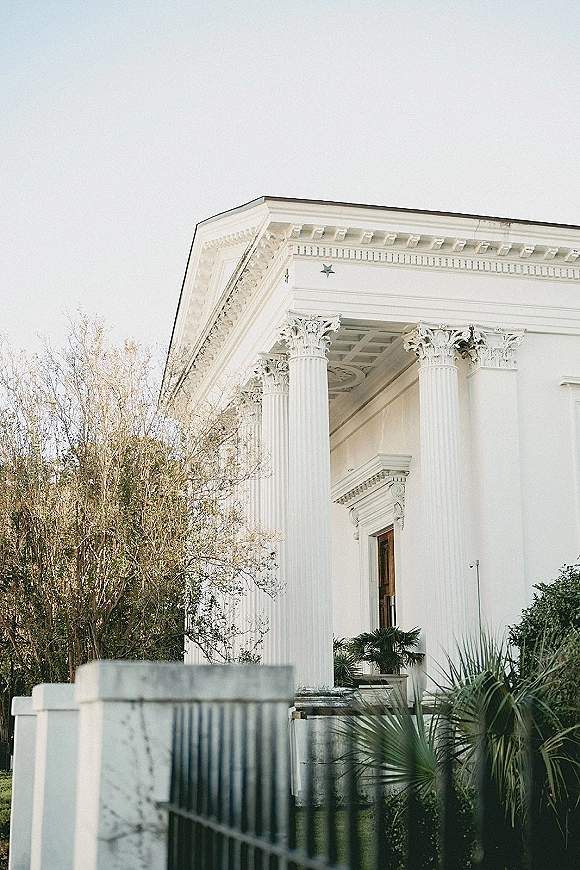 Wedding venue exterior with white column venue portico and pediment, framed by a wrought iron fence, palms, and manicured lawn