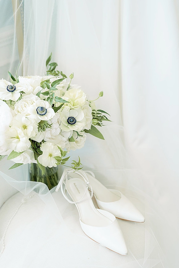 Bridal accessories flatlay with white bridal shoes and bouquet of white flowers and greenery, beside a tulle veil on soft white fabric in window light