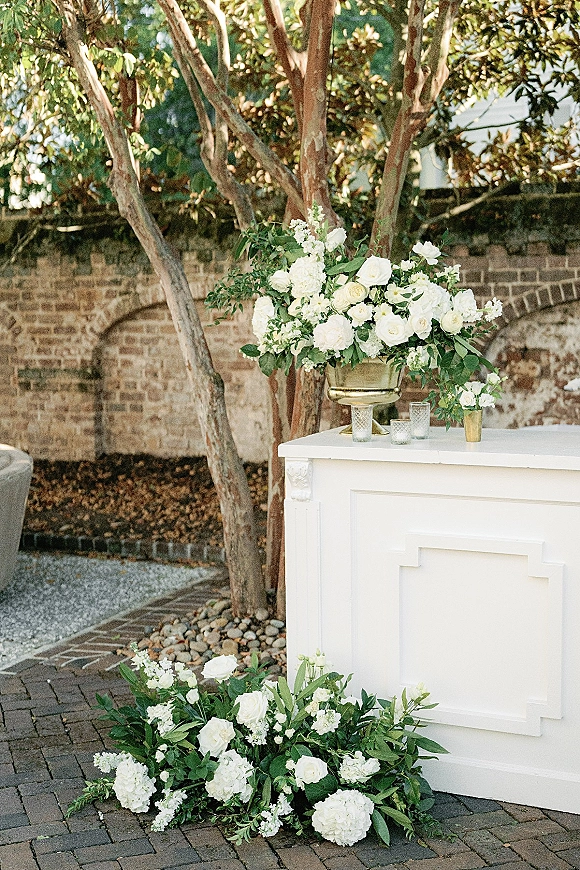 Wedding bar setup with a white wedding bar, lush white roses and hydrangeas in a brass urn, glass votives, and greenery by a brick wall