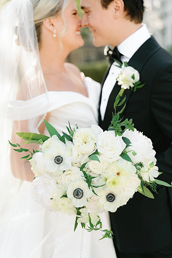 Wedding couple portrait of bride and groom close up sharing a nose kiss, bride holding white bouquet with greenery in a blurred garden backdrop
