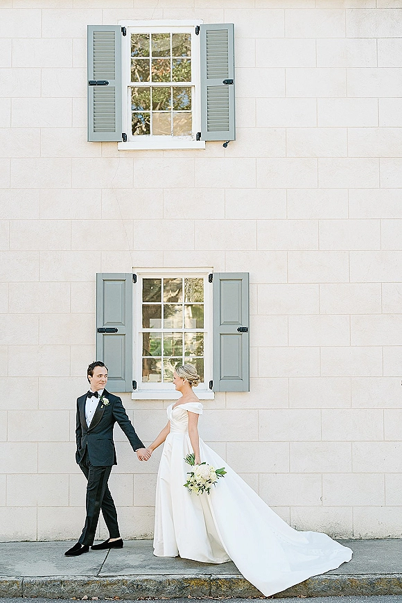Couple portrait of bride and groom holding hands, her long train and bouquet, his black tuxedo, by a white brick wall with shutters
