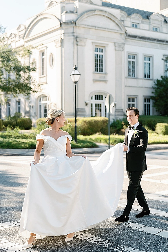 Couple portrait of bride and groom walking as he holds her dress train, crossing a crosswalk before a grand white estate facade in daylight
