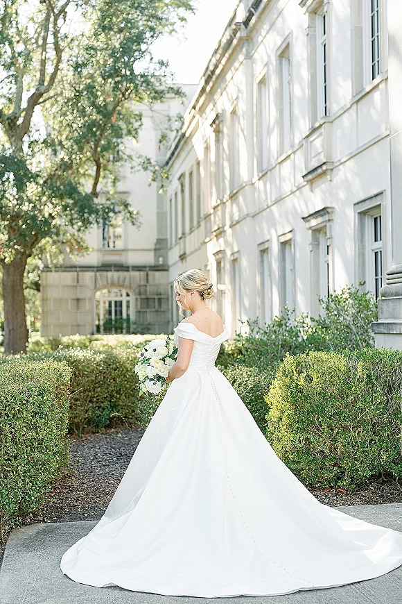 Bridal portrait of a bride in an off the shoulder wedding dress with long train, holding white rose bouquet in sunlight by a white facade