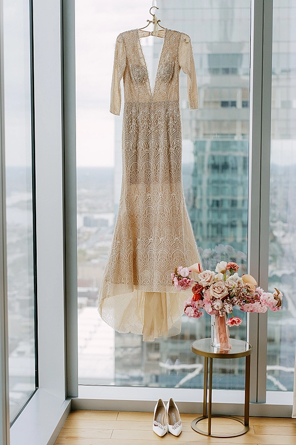 Wedding dress and lace wedding dress hanging by floor-to-ceiling windows, paired with jeweled heels and a rose bouquet on a side table.