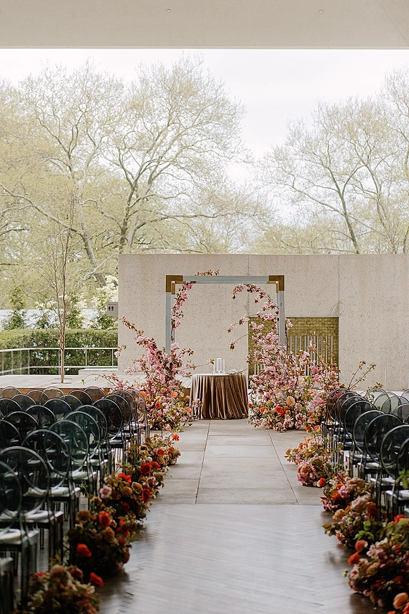 Ceremony setup with outdoor ceremony aisle lined in floral aisle arrangements, cherry blossom arch, clear chairs, and candles on a stone patio