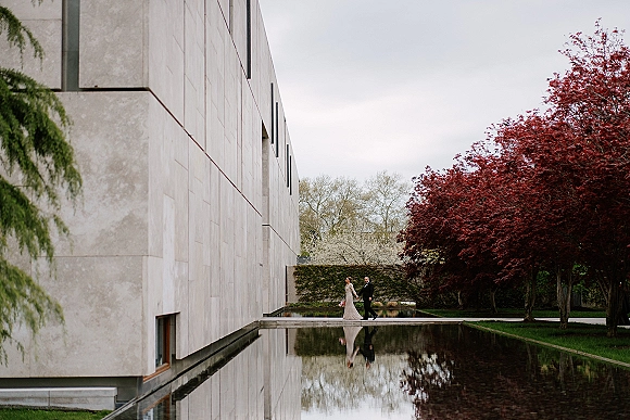 Couple portrait of bride and groom walking in a long sleeve wedding dress and tuxedo by a reflecting pool outside a modern stone building