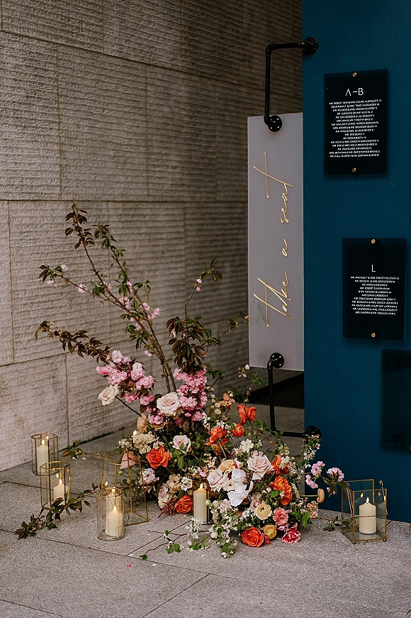 Wedding welcome sign acrylic with gold lettering, framed by cherry blossom florals and pillar candles in glass vases against a stone wall backdrop