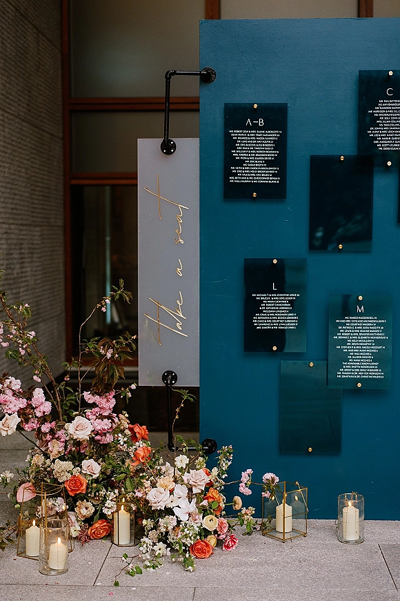 Seating chart display with an acrylic seating chart of clear cards on gold standoffs, set before a teal panel with roses and candle lanterns