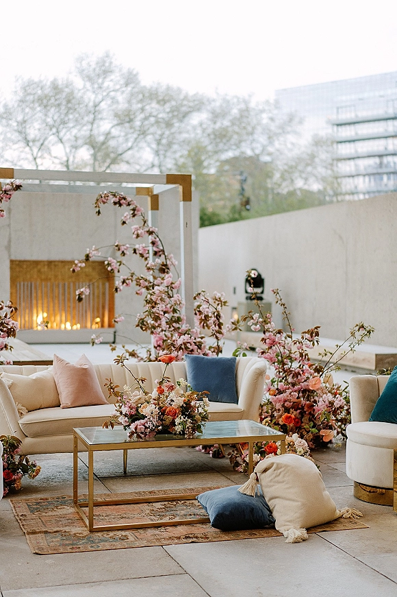 Wedding lounge setup with a cream velvet sofa, gold and glass coffee table, plush pillows, floral accents, and lanterns on a stone patio