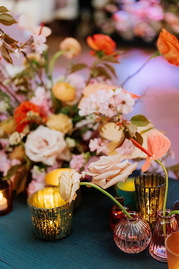 Reception tablescape with wedding table centerpiece of mixed roses and anthurium, greenery, and mercury glass votive candles under ambient lighting